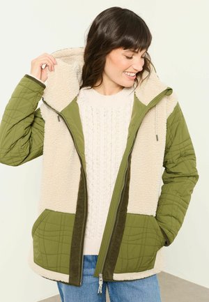 Kvinde smilende mens hun justerer hætten på en grøn quiltet og fleeceforet jakke over en hvid sweater og blå jeans.