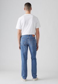 Homme debout, de dos, portant un t-shirt blanc, un jean bleu et des baskets blanches sur un fond gris clair.