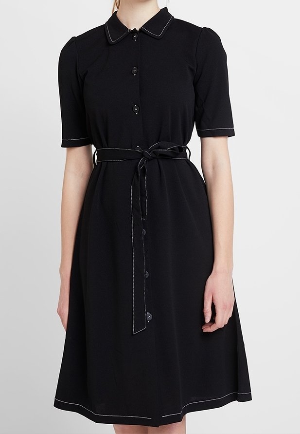 Femme portant une robe noire mi-longue à manches courtes, col, boutons noirs sur le devant et ceinture nouée à la taille.