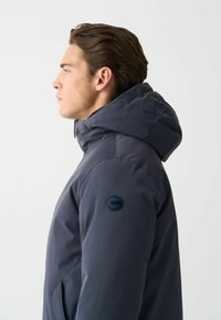 Muž v profile nosí tmavomodrú zimnú bundu s kapucňou, so zipsom na golieri a okrúhlym logom na hornej časti rukáva.