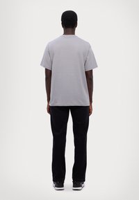 Uomo in piedi con le spalle rivolte, indossa una t-shirt a maniche corte grigio chiaro, pantaloni neri e sneakers bianco e nere, su uno sfondo neutro.