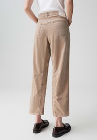 Pantaloni beige a gamba larga con vita alta, tasche posteriori e dettagli sulle cuciture, realizzati in un tessuto morbido. Abbinati a scarpe nere lucide.