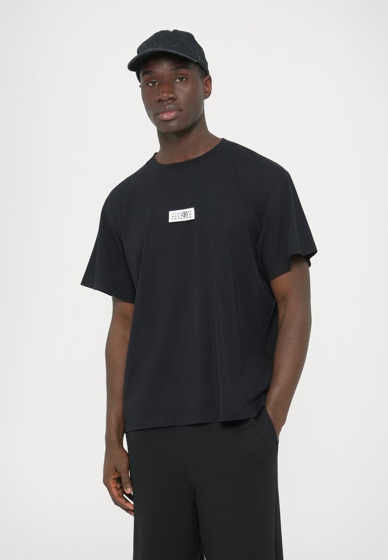 Sort bomulds-T-shirt med korte ærmer og afslappet pasform, med en hvid rektangulær logo-patch på brystet. Kombineret med sorte bukser.