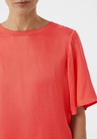 Koraalkleurig T-shirt met korte mouwen en een bootnek. De soepele, lichte stof heeft een subtiele glans en valt soepel om het lichaam.