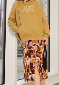 Beige hoodie with "AIR" logo, paired with a patterned skirt in brown, purple, and orange tones, featuring a side slit and button detailing.