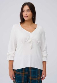 Witte lange mouwen top met een V-hals, gerimpelde details bij de buste en elastische manchetten, gedragen met een geruite broek.