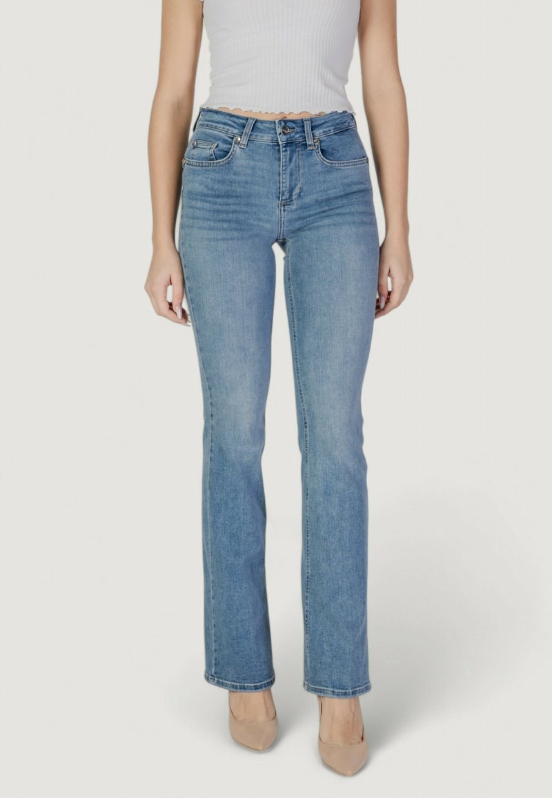 Femme portant un jean taille haute bleu clair à jambes droites, un haut court blanc côtelé, et des talons beiges à bouts pointus, debout sur un fond blanc.