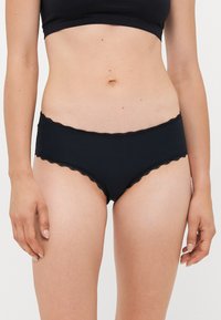 Kvinna som bär svarta ribbade hipsterunderkläder med spetsdetaljer, visad från midjan till övre låren mot en vit bakgrund.