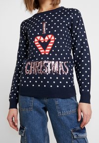 Pull marine avec des pois blancs, ornée d'un motif de cœur en canne de sucre rouge et blanc et d'un texte "J'aime Noël" en sequins. Poignets et ourlet côtelés.