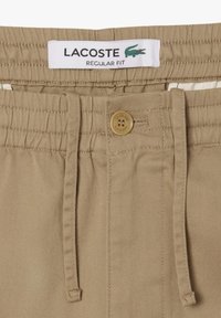 Beige regulære shorts med elastisk liv, snøring og knappelukking. Lacoste-logo på lappen ved livet.