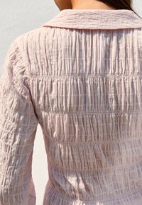Light pink garment with textured, gathered fabric. Features a classic collar and alternating horizontal seams for visual interest.
