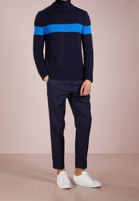Maglione a coste blu navy con collo alto e una larga striscia blu, abbinato a pantaloni tapered blu navy e sneakers bianche. Texture liscia, design aderente.