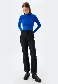 Blaues langärmliges Rollkragenoberteil kombiniert mit schwarzen schmal zulaufenden Hosen und schwarzen klobigen Stiefeln. Glatte Textur, minimalistisches Design, funktionale Taschen.
