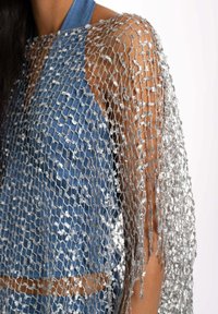 Capa de punto abierto en plateado sobre un top halter de denim, con textura de tejido suelto y bordes con flecos, que capta la luz con una calidad reflectante.