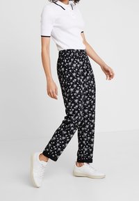 Pantalons noirs avec un motif de palmiers blancs, taille élastique, ourlets retroussés, associés à un polo blanc à manches courtes et des baskets blanches.