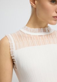 Hvid ærmeløs top med en ribbet tekstur, gennemsigtig panel ved halsen og delikate flæse detaljer langs kraven og ærmegabene.