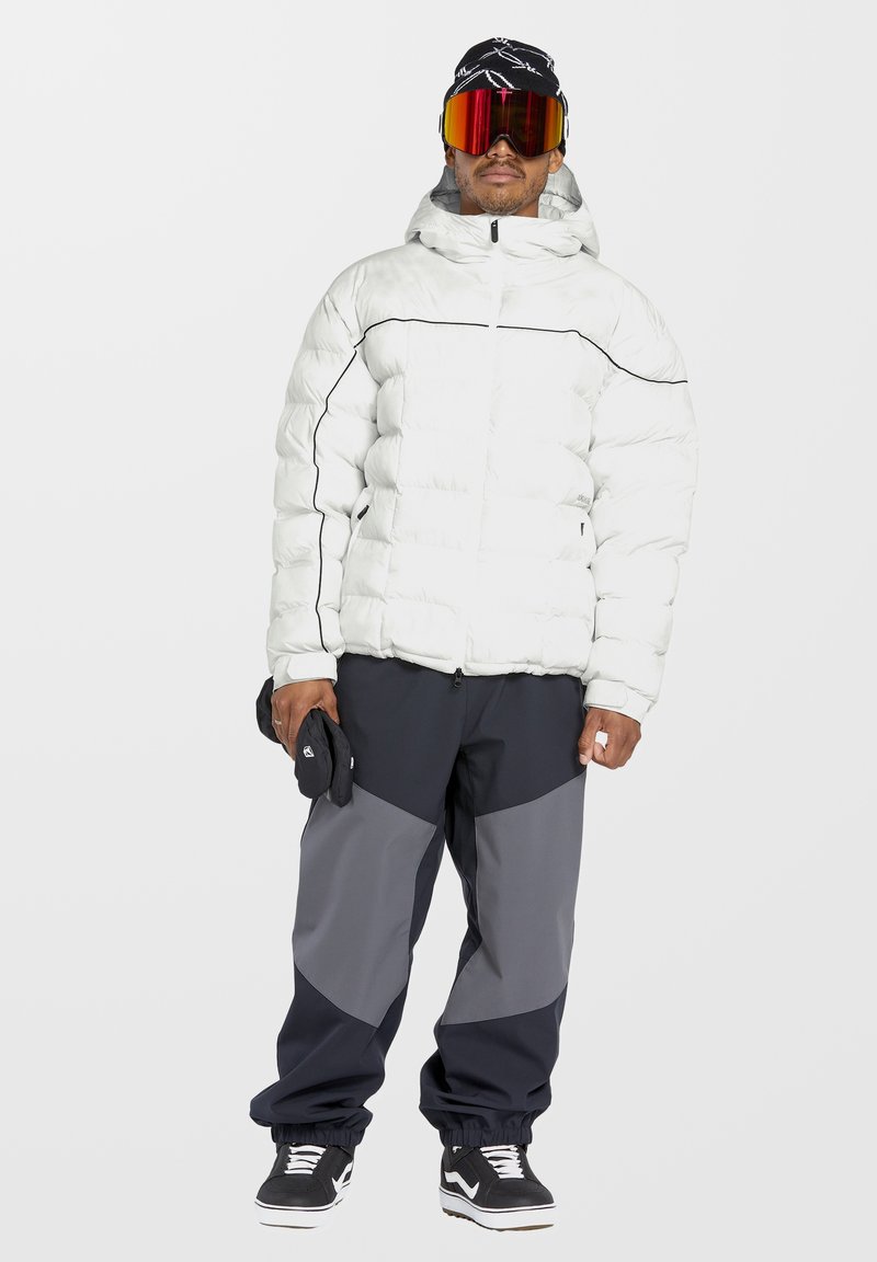 Wit pufferjack met zwarte accenten, rits aan de voorkant en capuchon. Gemanaged met marineblauwe en grijze skibroeken, zwarte sneakers en oranje spiegelgoggle.