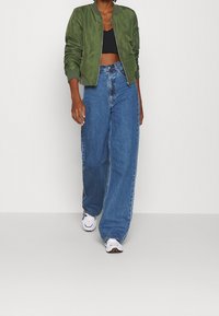 Zielona kurtka bomberka, czarny krótki top, luźne niebieskie dżinsy i białe sneakersy z kolorowymi akcentami. Strój charakteryzuje się swobodnym krojem i casualowym stylem.