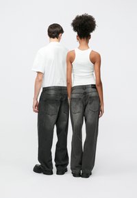 Dvema osebama stojita hrbt ob hrbtu, vsaka nosi prevelike črne jeans hlače z izrabljenim videzom in dvema zadnjima žepoma.