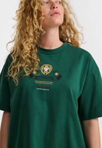 Zielona bawełniana koszulka T-shirt o luźnym kroju, z nadrukowaną grafiką w kolorze żółtym i czarnym, zawierającą logo marki i tekst, okrągły dekolt.
