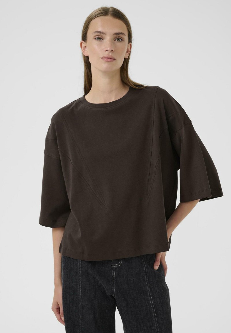 Braunes, übergroßes T-Shirt aus Baumwolle mit rundem Halsausschnitt, heruntergesetzten Schulternähten und kantigen Nahtt details. Getragen mit dunklen Jeans.