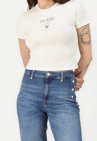 Camiseta ajustada en color blanco roto con manga corta y cuello redondo; presenta el logo "GUESS" en gris; jeans de mezclilla azul de tiro alto con detalles de botones.