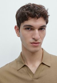 Jasnobrązowa koszulka polo o dzianinowej fakturze i dekolcie w serek. Model ma krótkie, kręcone włosy i neutralny wyraz twarzy. Czyste białe tło.