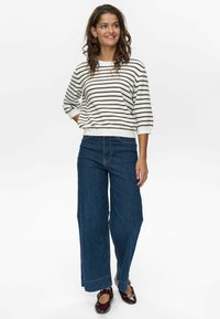 Randig vit och brun stickad tröja med lös passform, kombinerad med vida blå denimjeans med synlig sömnad och fållaccenter. Mörka skor.