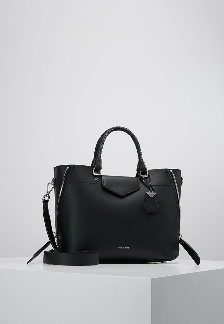 Sac à main en cuir noir avec poignées tressées, bandoulière détachable, détails de fermeture éclair argentés et petit rabat avant sur une surface blanche.
