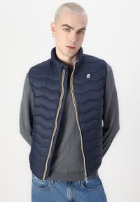 Gilet matelassé bleu marine avec un motif en chevrons, présentant un col montant, une fermeture éclair et un logo brodé sur la poitrine gauche.