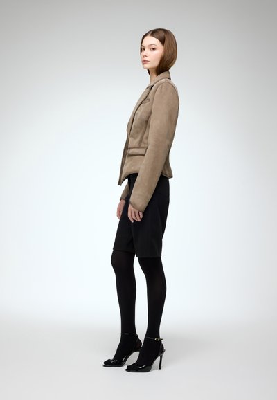 Beige suede jacket with a structured collar, paired with a black skirt. Model wears black tights and patent leather heels. Minimalist background.