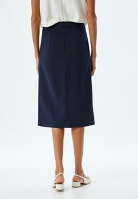 Navy-blauer Midirock mit geradem Design, der sanfte Falten an der Taille aufweist. Glatter Stoff und eine klassische Silhouette. Kombiniert mit weißen Schuhen.