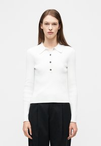 Γυναίκα που φοράει λευκό ριγωτό πόλο με μακριά μανίκια και τέσσερα κουμπιά, καθώς και μαύρο παντελόνι ψηλόμεσο με φαρδιά πόδια, στέκεται μπροστά σε απλό φόντο.