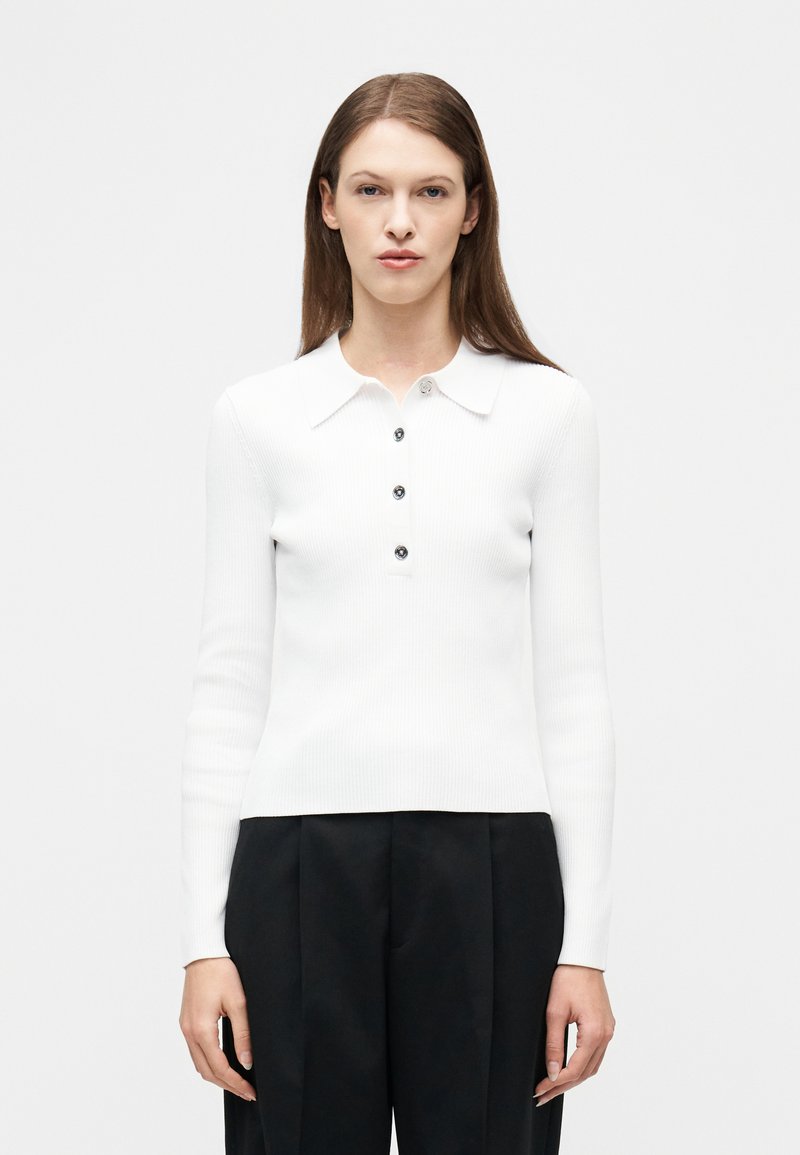 Γυναίκα που φοράει λευκό ριγωτό πόλο με μακριά μανίκια και τέσσερα κουμπιά, καθώς και μαύρο παντελόνι ψηλόμεσο με φαρδιά πόδια, στέκεται μπροστά σε απλό φόντο.