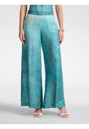 Pantalon large couleur sarcelle avec motif jacquard floral subtil, porté avec des talons ouverts blancs et des bracelets au poignet droit.