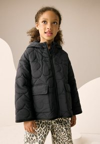 Veste noire matelassée avec capuche, ornée de coutures à motifs et de poches avant, associée à un pantalon imprimé léopard. Texture lisse.