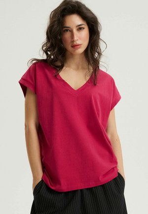 Młoda kobieta o falowanych ciemnych włosach, ubrana w luźną czerwoną bluzkę z krótkim rękawem i dekoltem w serek oraz czarne spodnie w paski, trzymająca ręce w kieszeniach, na neutralnym tle.