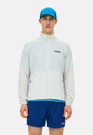 Junger Mann trägt eine hellgraue Jacke mit Reißverschluss, blaue Sportshorts und eine passende blaue Mütze, steht vor einem schlichten weißen Hintergrund.