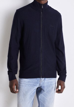Mand iført en mørk marineblå striktrøje med lynlås, ribstrikkede manchetter og kant, kombineret med lyseblå jeans, mod en ensfarvet hvid baggrund.