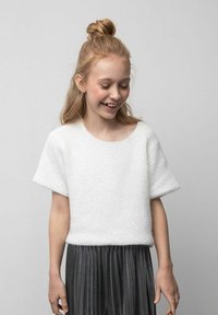 Biała, krótka, teksturowana dzianinowa bluzka z krótkim rękawem w zestawieniu z plisowaną, ciemnoszarą spódnicą. Strój ma okrągły dekolt i luźny krój.