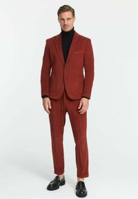 Roter Cordanzug mit strukturiertem Finish, bestehend aus einem einreihigen Blazer und schmal geschnittenen Hosen, kombiniert mit einem schwarzen Rollkragenpullover und Lederschuhen.