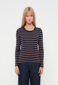 Femme portant une chemise à rayures horizontales bleu marine et marron à manches longues et un jean large foncé, se tenant devant un fond blanc.