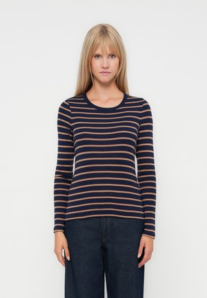 Woman wearing a navy and brown horizontal striped long-sleeve shirt and dark wide-leg jeans standing against a white background.