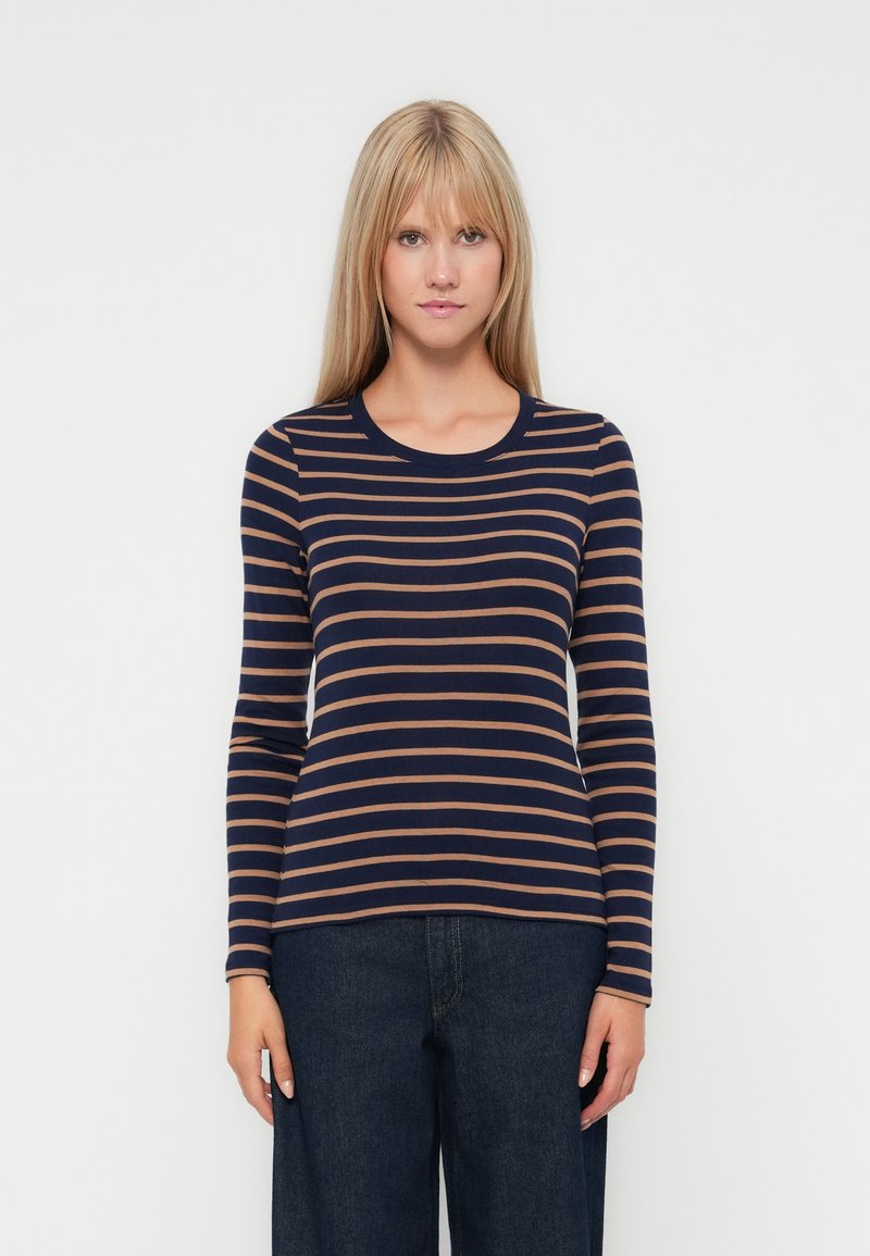 Femme portant une chemise à rayures horizontales bleu marine et marron à manches longues et un jean large foncé, se tenant devant un fond blanc.