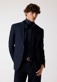 Marineblauw blazer met een getailleerde pasvorm, met een notch revers, sluiting met twee knopen en een subtiele textuur, gecombineerd met een donkere coltrui en denim jeans.