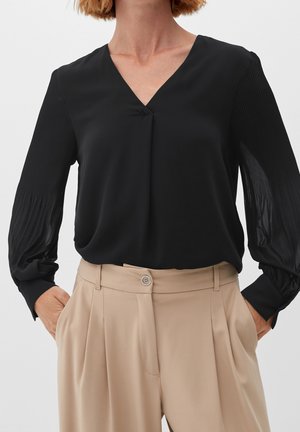Kvinde iført en sort langærmet V-hals bluse med plisserede ærmer og beige højtaljede plisserede bukser med hænderne i lommerne.