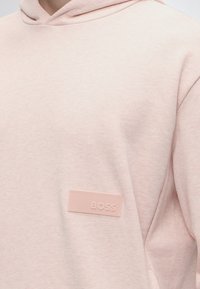 Sweat à capuche rose clair avec un petit patch rectangulaire près de la poitrine portant le mot « BOSS » en relief dans le même rose.