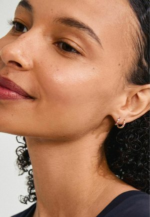 Goldener Ohrring mit bunten Edelsteinen, mit einem gedrehten Design. Nahaufnahme eines Gesichts mit lockigem Haar und glatter Haut.