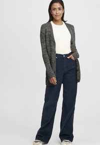 Long cardigan gris avec texture côtelée, doté de boutons en bois, associé à un pantalon en velours côtelé bleu marine taille haute. Haut de couleur claire en dessous.