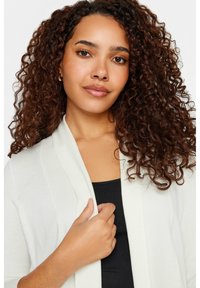 Cardigan ligero de frente abierto en suave tejido blanco con textura acanalada. Presenta mangas largas y un ajuste relajado, llevado sobre una camiseta negra.